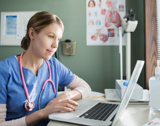 A woman wearing scrubs sits at a desk in a hospital setting and uses a laptop computer.